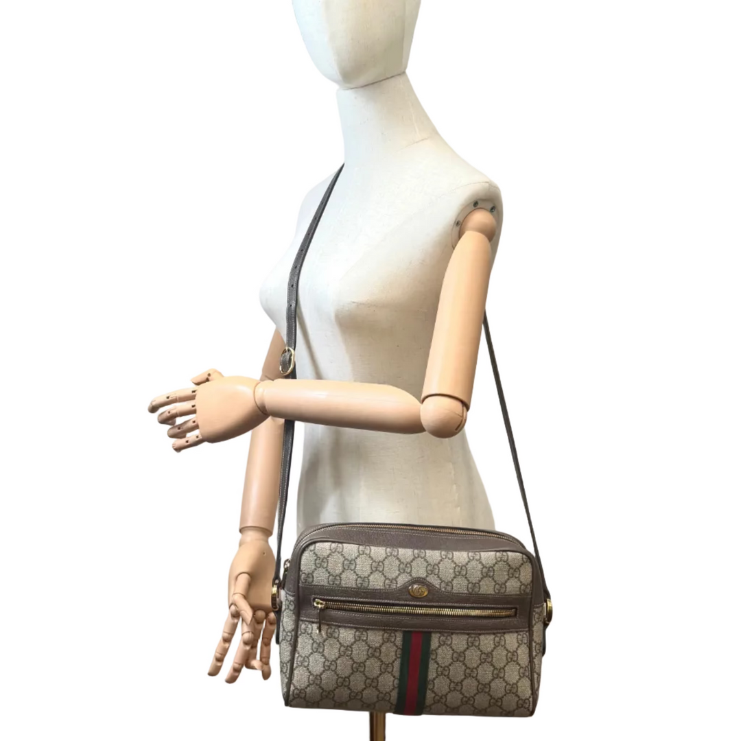 A Gucci shoulder bag with the brand's classic GG monogram pattern and green-red-green stripe, displayed on a wooden articulated mannequin torso in a room with a white wall, painting, and studio equipment.