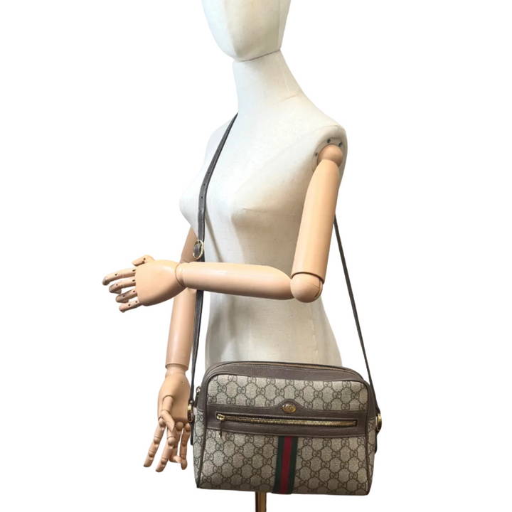 A Gucci shoulder bag with the brand's classic GG monogram pattern and green-red-green stripe, displayed on a wooden articulated mannequin torso in a room with a white wall, painting, and studio equipment.