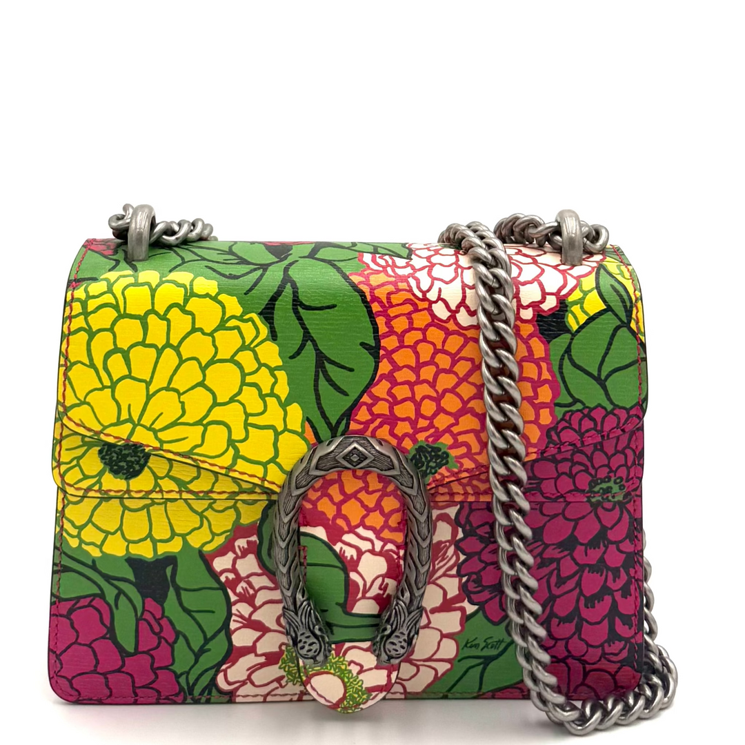 A small structured handbag with a vibrant multicolor floral and fruit print, featuring a silver-tone chain strap and a central metal logo clasp, displayed against a white studio backdrop.