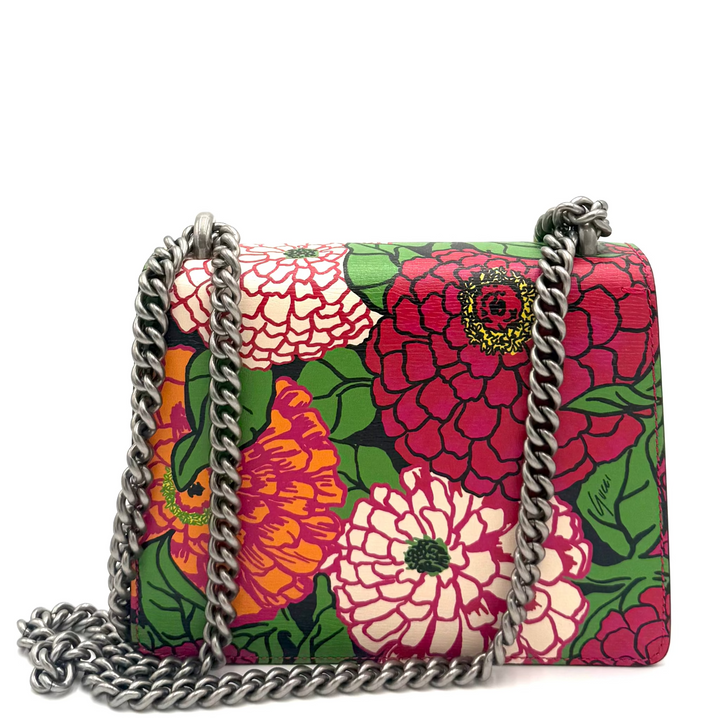 A small rectangular shoulder bag with a vibrant floral print featuring pink, red, orange, and white flowers with green leaves, accented by a silver-tone chain strap, set against a plain white background.