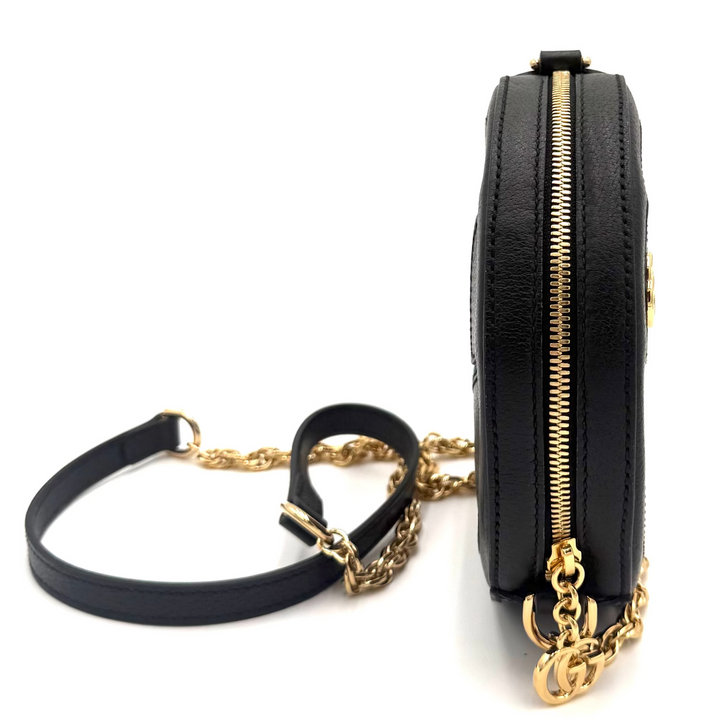 A black leather crossbody bag with a gold chain strap and gold zipper, shown from a side profile view on a white background.