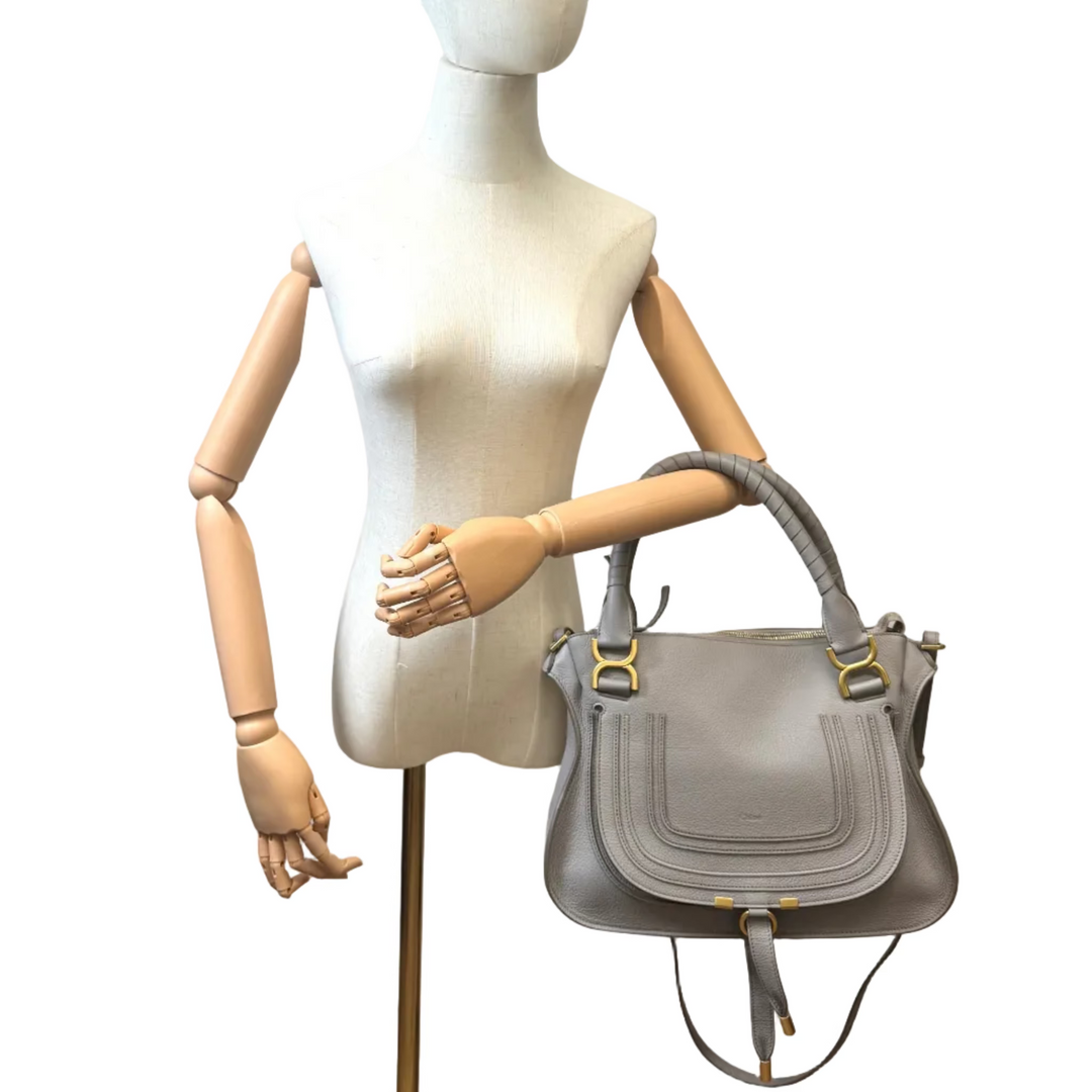A light gray leather handbag with gold-tone hardware, displayed on a beige wooden articulated mannequin against a plain white background.