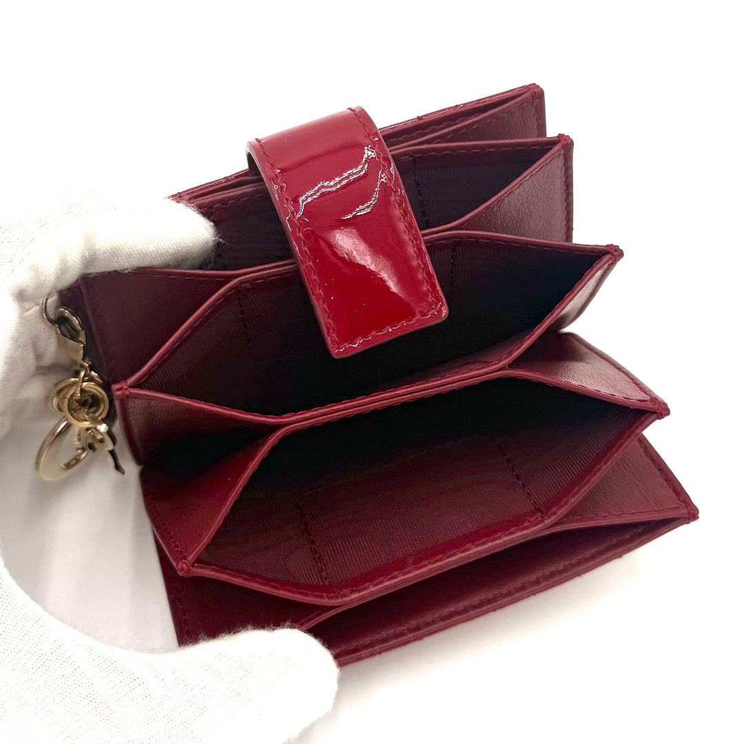 A burgundy patent leather multi-card wallet held open by a white gloved hand, showing its interior compartments and gold-tone zipper pull against a white background.