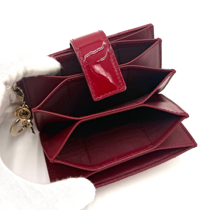 A burgundy patent leather multi-card wallet held open by a white gloved hand, showing its interior compartments and gold-tone zipper pull against a white background.