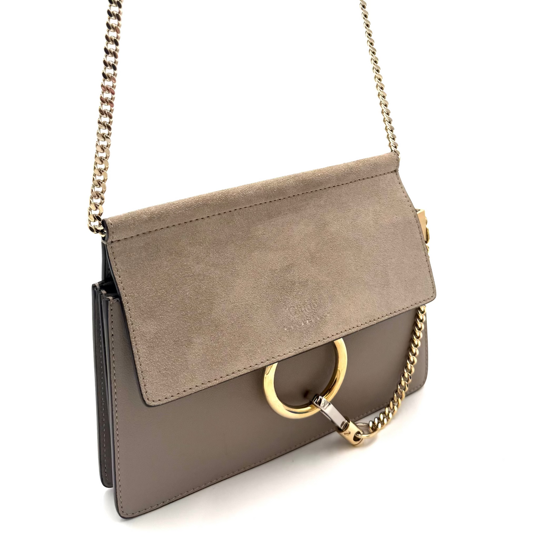 A taupe-brown leather shoulder bag with a suede flap, gold-tone chain strap, and a large circular gold ring detail on the front, shown against a plain white background.