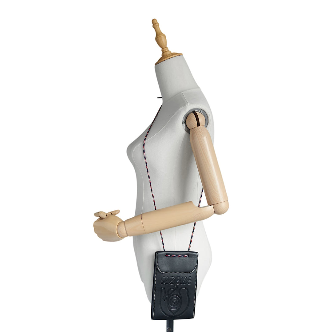 A black leather crossbody pouch with a circular logo, shown hanging from a wooden mannequin torso against a plain white background.