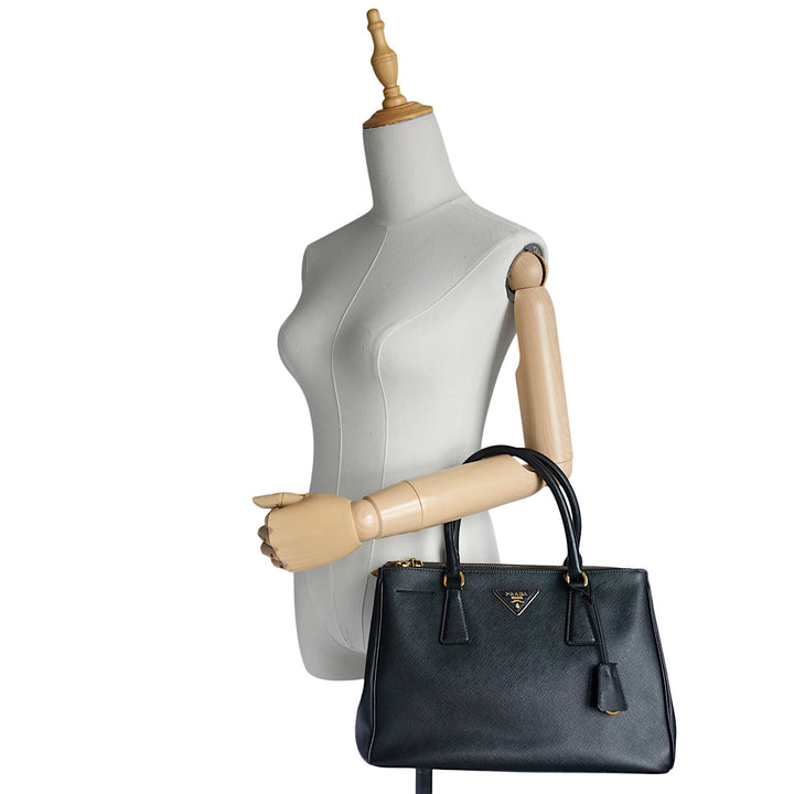 A structured black leather handbag with gold hardware and a triangular Prada logo plaque, displayed on a light gray mannequin torso with wooden arms against a white background.
