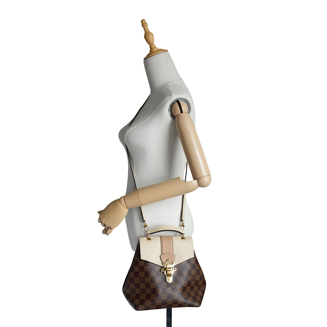 A Louis Vuitton shoulder bag in brown Damier canvas with a beige leather flap and trim, displayed on a white mannequin torso with wooden arms.