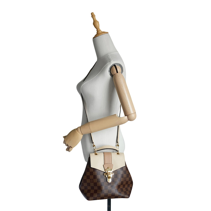 A Louis Vuitton shoulder bag in brown Damier canvas with a beige leather flap and trim, displayed on a white mannequin torso with wooden arms.